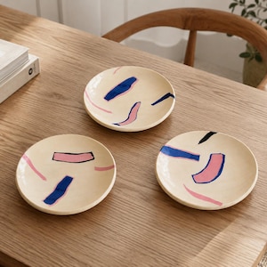 May include: Three round ceramic plates with a cream-coloured base, each featuring a unique arrangement of abstract brushstrokes in shades of navy blue, light pink, and black. The plates have a smooth, glossy finish and a simple, circular shape.