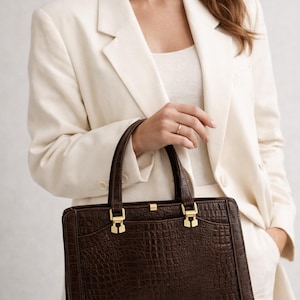 May include: A dark brown, rectangular handbag with a crocodile skin texture. The bag has two handles and gold-colored clasps. The bag is being held by a person wearing a cream-colored blazer and pants.