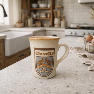 May include: A speckled beige ceramic mug featuring a brown and blue shield-shaped emblem on the front. The emblem displays the text Clovelly above illustrations of two donkeys and three fish. The mug has a sturdy handle and a smooth rim.