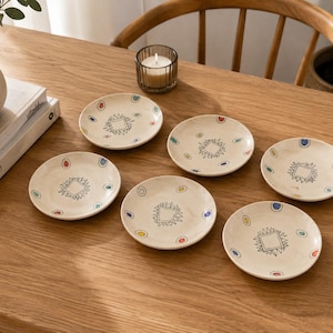 May include: A set of six round, off-white ceramic plates, each featuring a central black line-drawn geometric square motif surrounded by small, colourful painted oval accents in shades of blue, red, yellow, and green along the rim.