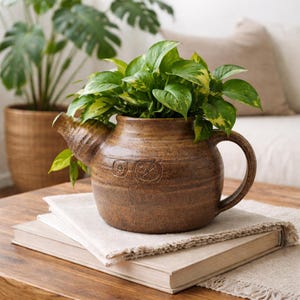 Puede incluir: Una maceta de cerámica marrón con forma de tetera llena de plantas de potos verdes y amarillas. La maceta está sobre una pila de libros y un paño de lino. Otra planta en maceta es visible en el fondo.