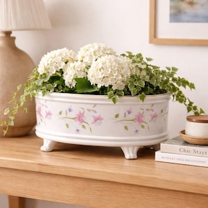 Puede incluir: Una jardinera de cerámica blanca con un diseño floral, llena de hortensias blancas y follaje verde. La jardinera ovalada tiene pequeños pies y un borde dorado. El patrón floral incluye flores rosas y moradas.