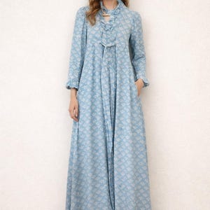 May include: A long, light blue dress with a floral pattern. The dress features long sleeves, a ruffled collar, and a tie detail at the neckline. The dress has a flowing skirt and pockets.