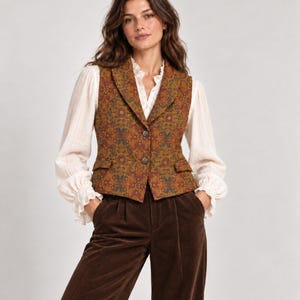 May include: A sleeveless waistcoat featuring a dark orange and green floral tapestry pattern with two front buttons and two small flap pockets. The waistcoat is worn over a long-sleeved white blouse with ruffled cuffs and paired with dark brown corduroy trousers.