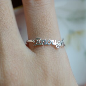 May include: A silver ring with the word "Enough" in a cursive font. The ring is thin and delicate, with the word spanning across the top. The ring is shown on a finger, with a blurred background.