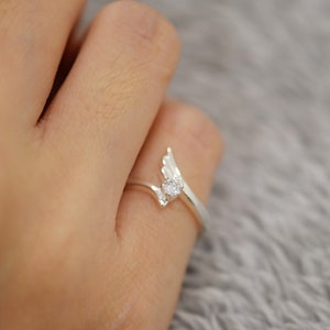 Puede incluir: Un anillo de plata con un delicado diseño de ala, con una pequeña y brillante gema en el centro. La banda del anillo se curva con gracia, creando una estética elegante y minimalista. El anillo se muestra en una mano.