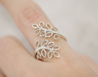 May include: A silver-colored ring featuring a leaf design. The ring wraps around the finger with two leaf clusters, creating an open, adjustable style. The leaves have detailed vein patterns, adding a naturalistic touch to the jewelry.