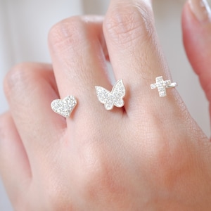 Puede incluir: Tres anillos de plata con diferentes diseños: un corazón, una mariposa y una cruz, todos incrustados con pequeñas piedras brillantes. Los anillos se muestran en un dedo, mostrando los delicados detalles y el brillo de la joyería.