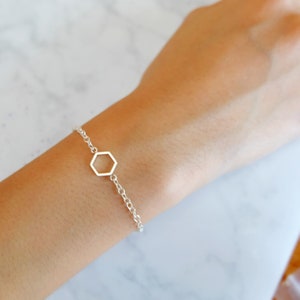 May include: A silver bracelet featuring a delicate chain and a geometric hexagon charm. The bracelet is shown on a wrist, showcasing its simple yet elegant design. The bracelet's length is approximately 7 inches.
