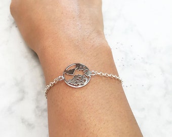May include: A silver bracelet featuring a circular charm with a cut-out design of a landmass and ocean. The bracelet is on a chain and is worn on a wrist. The jewelry is made of a shiny metal.
