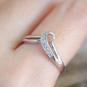 Op de afbeelding: Een zilveren ring met een gebogen ontwerp, bezet met kleine, lichtblauwe edelstenen. De ring heeft een delicate, elegante uitstraling, met een gepolijste afwerking. De edelstenen zijn in een rij langs de boog van de ring gerangschikt.