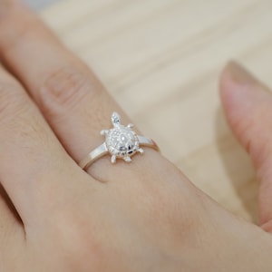 May include: A silver ring featuring a detailed turtle design. The turtle has a textured shell and is the focal point of the ring. The ring is shown on a finger, highlighting the delicate craftsmanship and the ring's size.