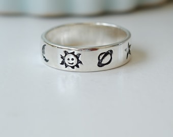 May include: A silver ring with etched celestial designs. The ring features a smiling sun, a crescent moon, a planet with rings, and a star. The ring is on a white surface.