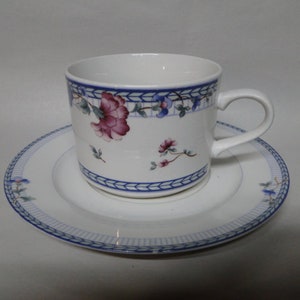 Może przedstawiać: Zestaw filiżanki i spodeczka z białej porcelany z niebieskim i kwiatowym wzorem. Filiżanka ma uchwyt, a spodeczek ma falisty brzeg.