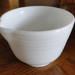 May include: A white ceramic mixing bowl with a pouring spout. The bowl has a smooth, glossy finish and features three raised bands around the exterior.