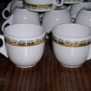 May include: White ceramic coffee cups with a gold and black geometric pattern around the rim. The cups have a rounded shape with a handle. Several cups are stacked in the background, with two in focus.