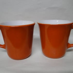 Puede incluir: Dos tazas de color naranja con el interior blanco. Las tazas tienen una forma redondeada y un asa en el lado derecho.