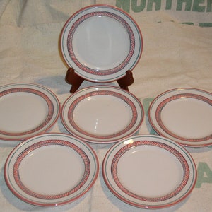 May include: A set of six white ceramic plates. Each plate features a red and blue rope-like border design. One plate is displayed on a wooden stand, while the other five are arranged on a white surface.