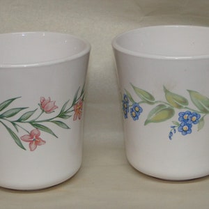 May include: Two white ceramic mugs with floral designs. One mug has pink and orange flowers, the other has blue flowers.