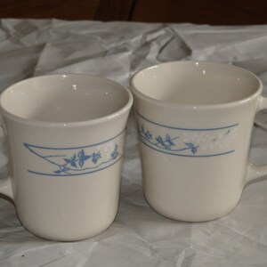 May include: Two white ceramic mugs with blue floral designs. The mugs have a handle on the right side.