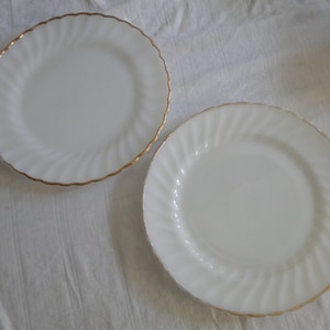 Puede incluir: Dos platos de cerámica blanca con un borde dorado y un diseño de espiral. Los platos están sobre una superficie blanca.