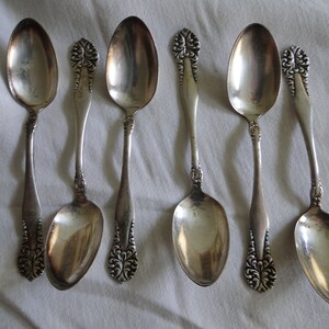 May include: Six antique silver spoons with ornate floral designs on the handles. The spoons are arranged on a white surface, showcasing their polished, reflective surfaces. The spoons vary slightly in shape and size, adding to their vintage charm.