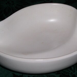 May include: A white ceramic bowl with a unique, asymmetrical shape. The bowl has a smooth, glossy finish and is slightly discolored in spots.
