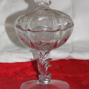 May include: A clear glass candy dish with a scalloped edge and a floral design. The dish has a footed base with a decorative swirl design and a lid with a floral finial.