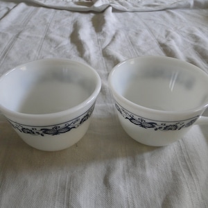 Puede incluir: Dos tazas de cerámica blanca con diseños florales azules. Las tazas tienen una forma redondeada y un asa en el lateral.