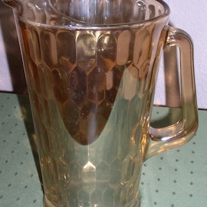 Puede incluir: Una jarra de vidrio ámbar transparente con un patrón de panal texturizado. La jarra tiene un asa y un pico.