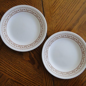 May include: Two white ceramic dinner plates with a brown floral pattern around the edge.