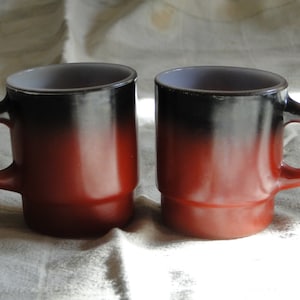Puede incluir: Dos tazas de cerámica con doble pared, con un degradado de negro a rojo y un interior blanco. Las tazas tienen un asa redondeada y una base ancha.