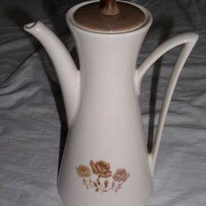 May include: Cream-colored ceramic coffee pot with a brown lid and handle. The pot features a floral design of brown roses. The handle is curved, and the spout is slender. A vintage-style coffee server.
