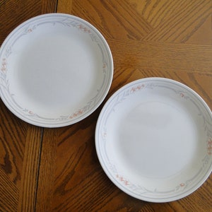 Könnte beinhalten: Zwei weiße Keramik-Dinnerteller mit einem Blumendesign in Grau- und Rosatönen. Die Teller haben einen weißen Hintergrund mit einem grauen und rosafarbenen Blumenrand.