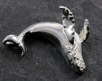 May include: A silver charm in the shape of a humpback whale, with detailed fins and a textured body. The whale is leaping out of the water.