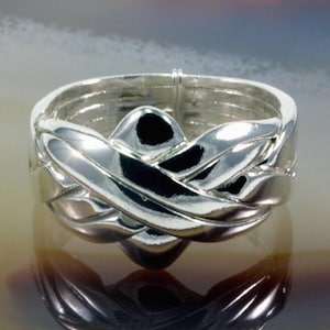 Könnte beinhalten: Ein silberner Puzzle-Ring mit einem komplizierten, ineinander verwobenen Design. Die Bänder des Rings sind glatt geschwungen und greifen ineinander, wodurch ein komplexes Muster entsteht. Das polierte Metall reflektiert das Licht und unterstreicht die detaillierte Handwerkskunst und den einzigartigen Stil des Rings.