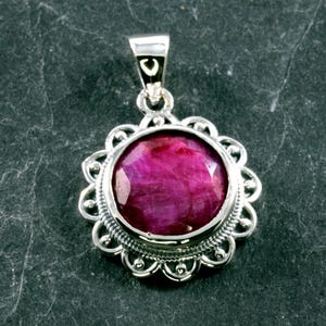 Könnte beinhalten: Silberanhänger mit einem großen, facettierten, runden Rubin-Edelstein. Der Rubin hat eine tiefrosa-rote Farbe und ist in einen dekorativen, silbernen Rahmen mit Muschelrand gefasst. Der Anhänger hat oben eine kleine Schlaufe für eine Kette.