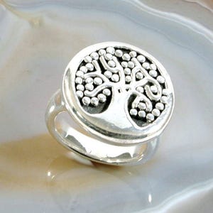 May include: A silver ring featuring a tree of life design. The ring is round and has a detailed design of a tree with branches and leaves.