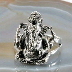 Könnte beinhalten: Ein silberner Ring mit einer detaillierten Darstellung der hinduistischen Gottheit Ganesha in sitzender Pose. Der Ring zeigt aufwendige Schnitzereien des elefantenköpfigen Gottes mit poliertem Finish. Der Ring ist ein religiöses Schmuckstück.