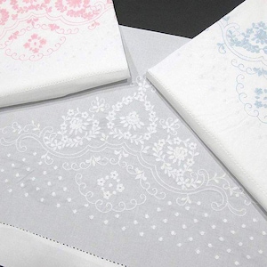 Puede incluir: Servilletas de lino blanco con delicados bordados florales en rosa y azul. Las servilletas están dobladas y apiladas, con un camino de mesa bordado a juego en primer plano. El diseño presenta patrones florales y de puntos.