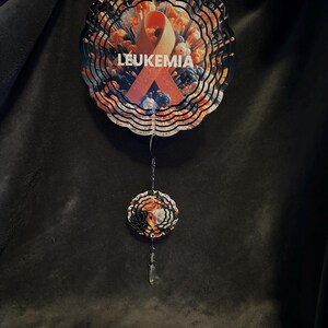 May include: A decorative wind spinner with a floral design and the word "LEUKEMIA" printed on it. The spinner features an orange ribbon, a symbol of leukemia awareness. The design includes orange, white, and blue colours.