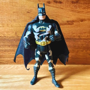 May include: A Batman action figure in a standing pose. The figure is predominantly black with white and gray accents, a yellow utility belt, and a black cape. The Batman logo is on the chest. The figure is approximately 6 inches tall.