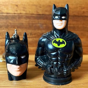 May include: Black Batman figurine and keychain. The figurine features a black suit with a yellow and green bat symbol. The keychain is a Batman head. Both items have a tan face and white eyes.
