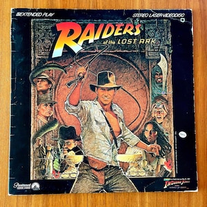 May include: A vintage "Raiders of the Lost Ark" stereo laser videodisc. The cover features a movie poster with Harrison Ford as Indiana Jones, holding a whip. The title is in large yellow and red letters.
