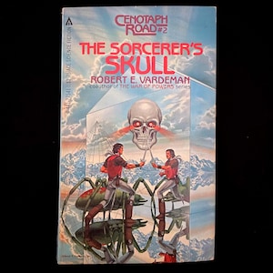 May include: A vintage paperback book titled "The Sorcerer's Skull" by Robert E. Vardeman. The cover art features a skull with glowing red eyes, two figures with swords, and a surreal landscape with mountains and a mirrored structure. The book is part of the Cenotaph Road series.