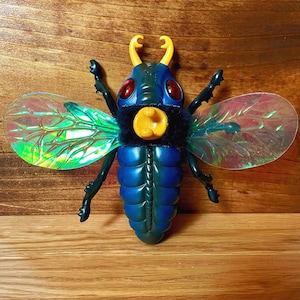 May include: A blue and black toy insect with iridescent wings, red eyes, and yellow antennae. The toy has a textured body and a fuzzy chest. The wings are translucent with green and purple hues. The insect is posed on a wooden surface.