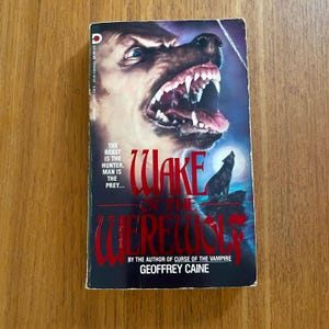 May include: A vintage paperback book titled "Wake of the Werewolf" by Geoffrey Caine. The cover features a snarling wolf head, a howling wolf silhouette, and the text "The Beast is the Hunter. Man is the Prey..."