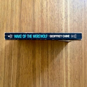 May include: A book titled "Wake of the Werewolf" by Geoffrey Caine. The spine is dark blue with white and teal lettering. The book is lying on a wooden surface.