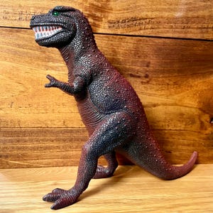 May include: A maroon and black Tyrannosaurus Rex toy dinosaur with green eyes and white teeth. The toy is made of a textured material and is posed in a standing position. The dinosaur is on a wooden surface.