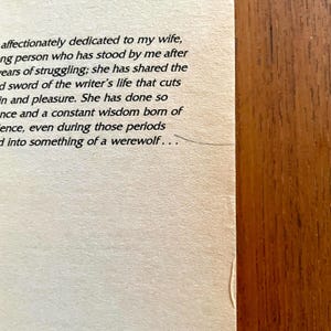 May include: Close-up of a book page with printed text. The text is in black font on a cream-colored paper. The right side of the image shows a wooden surface. The text includes phrases like "dedicated to my wife" and "something of a werewolf."